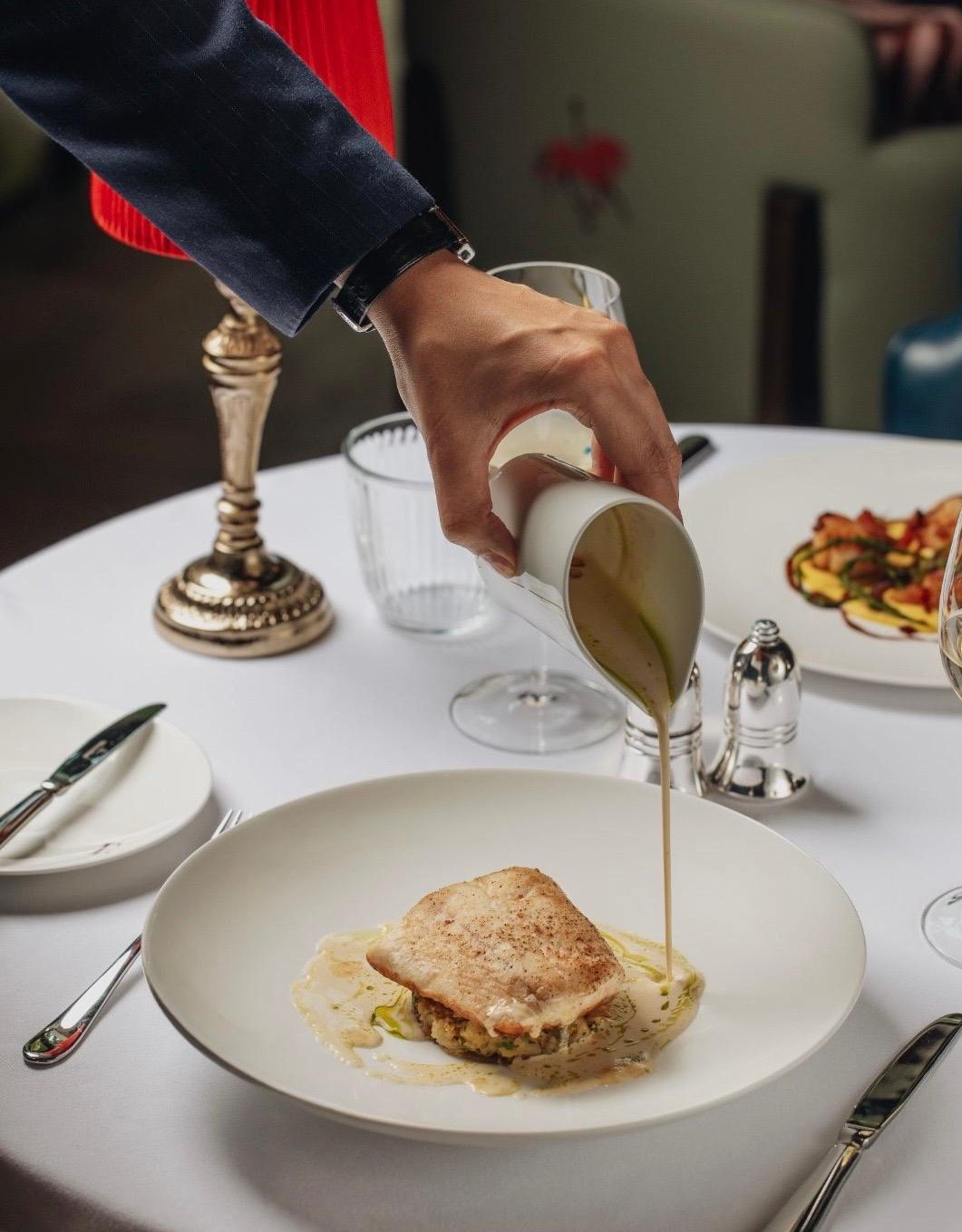 Waiter pouring cream sauce from a jug over a fish fillet on a white plate at a fine dining restaurant table.