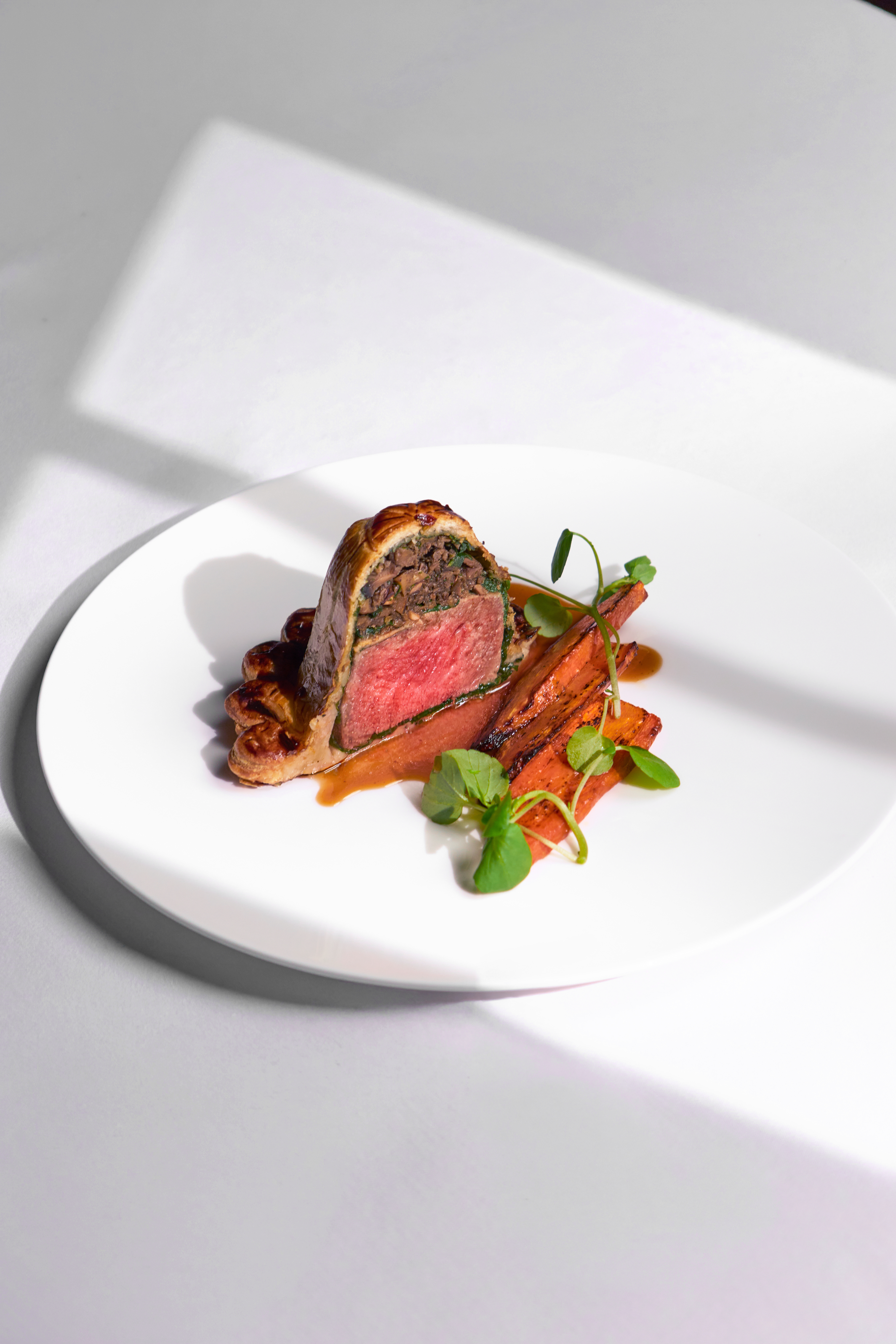 Sliced beef Wellington with pink medium-rare centre, roasted carrots, mushrooms, jus and micro herbs on a white plate