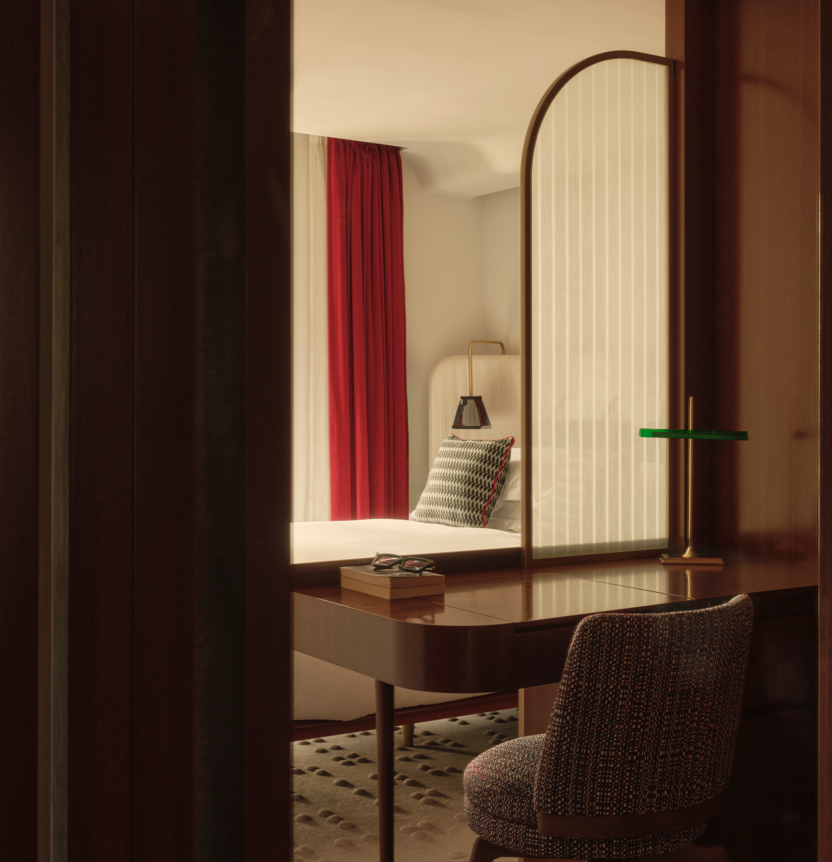 Hotel room with walnut desk, arched brass mirror, green desk lamp, textured chair, patterned floor, and red curtains visible in reflection.