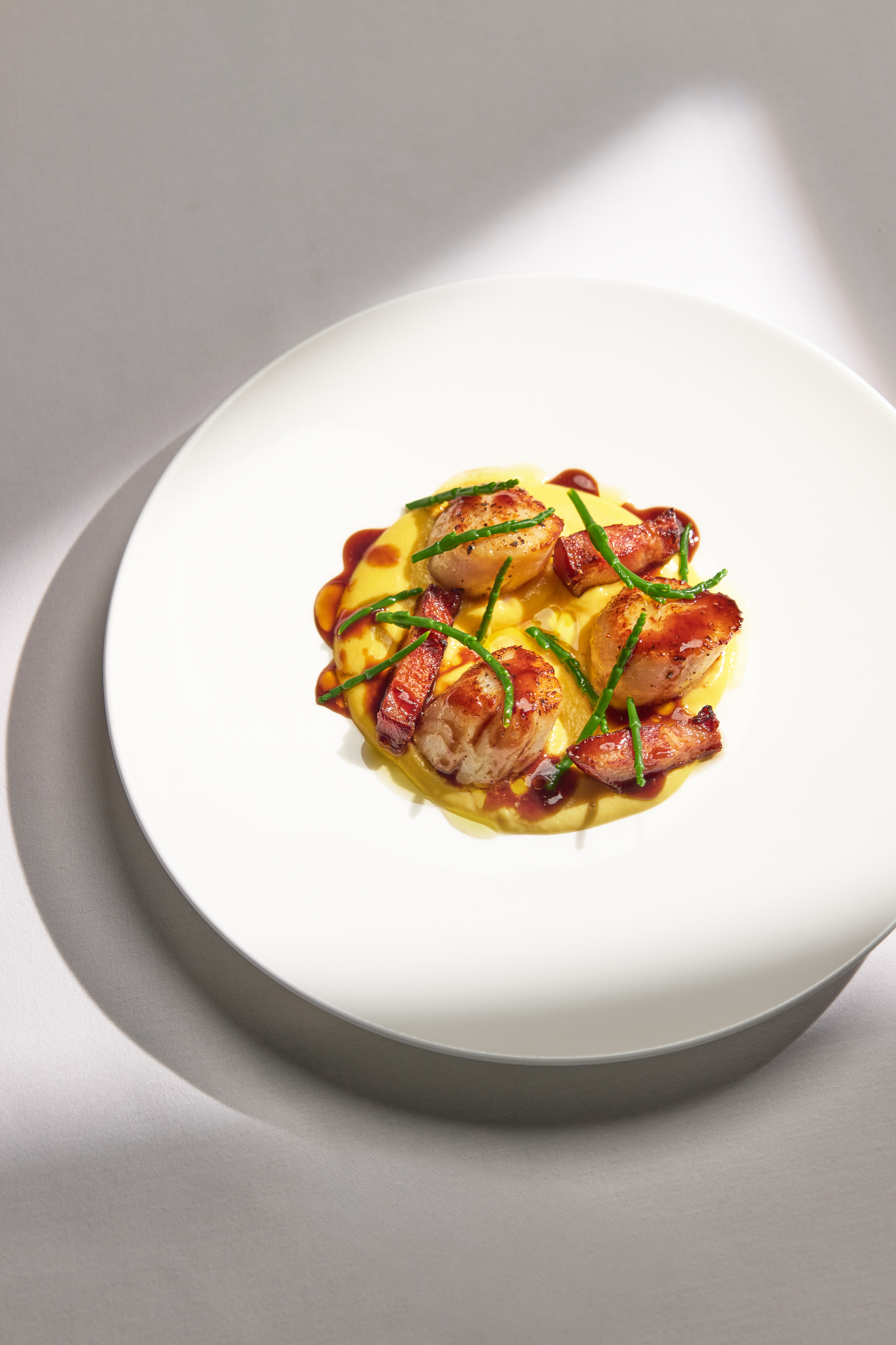 Seared scallops with crispy bacon on a yellow purée, garnished with samphire and a red sauce reduction, served on a round white plate.