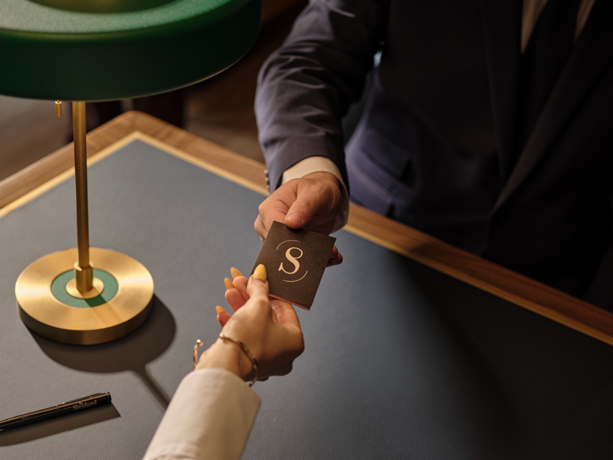 Hotel staff handing a dark brown key card with a gold 'S' logo to a guest at a reception desk with a brass lamp.
