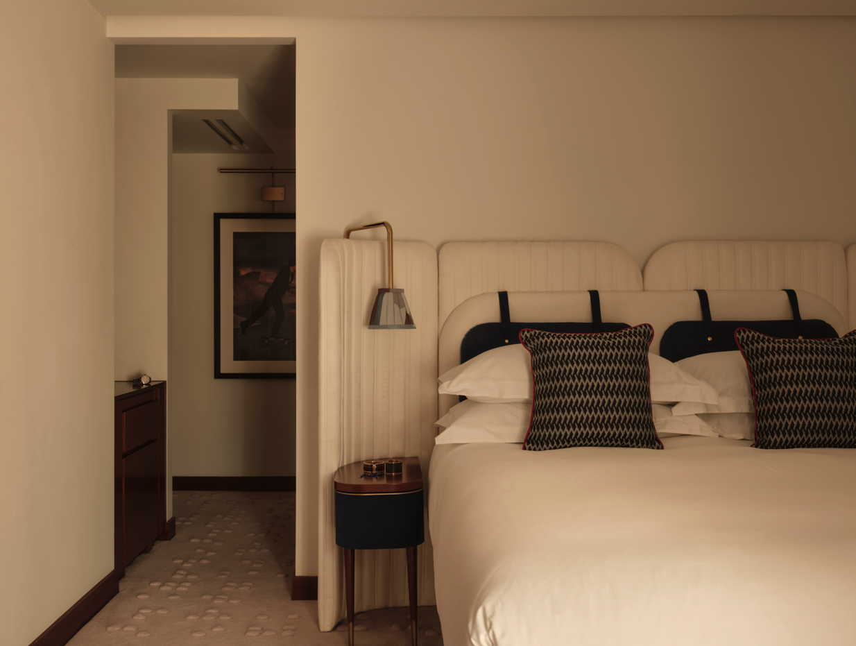 Hotel room with cream upholstered headboard, white bedding, dark patterned cushions, brass wall lamp, and a doorway leading to an adjoining room.