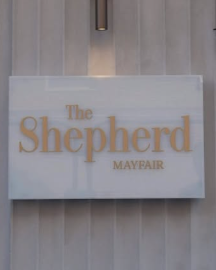 White rectangular sign with gold text reading 'The Shepherd Mayfair', mounted on a striped grey wall.