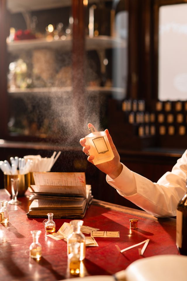Hand spraying a perfume bottle above a red desk scattered with small fragrance vials and cards, in a warmly lit perfumery.