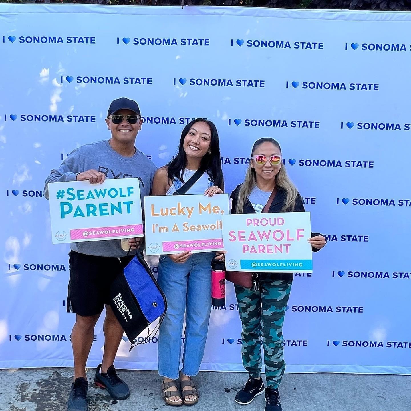 Three people pose holding signs in front of a backdrop that says Sonoma State University