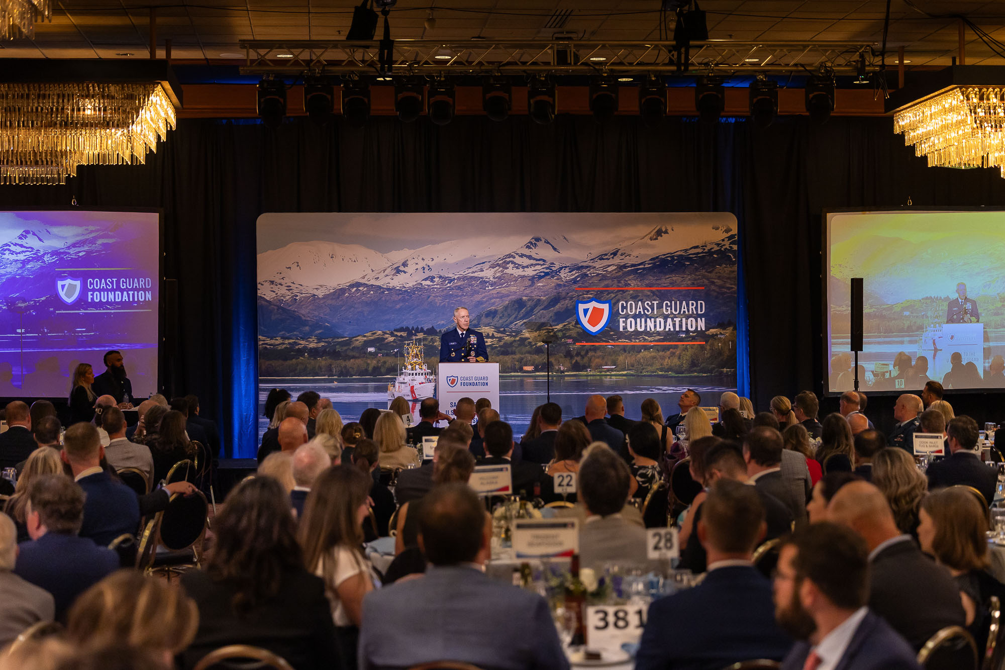 Crown seated in a large banquet hall as the Commandant of the U.S. Coast Guard delivers his address.