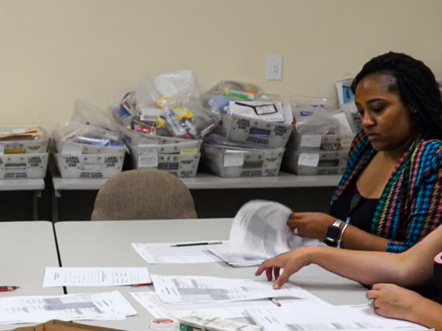 A woman with dark locks pulled back, in a bright teal, pink, and yellow woven jacket, looks down as she sorts papers at a long table; stacked gray postal trays fill the wall behind her. She sits at a light gray table covered in printed forms and pencils, lifting a sheet of paper with one hand. Another person's hands rest on the table in the foreground. Behind them, stacks of gray U.S. Postal Service trays hold clear plastic bags of small items, and a tan office chair sits in front of them against a cream wall. The scene suggests careful, hands-on work, such as sorting or reconciling ballots and records.