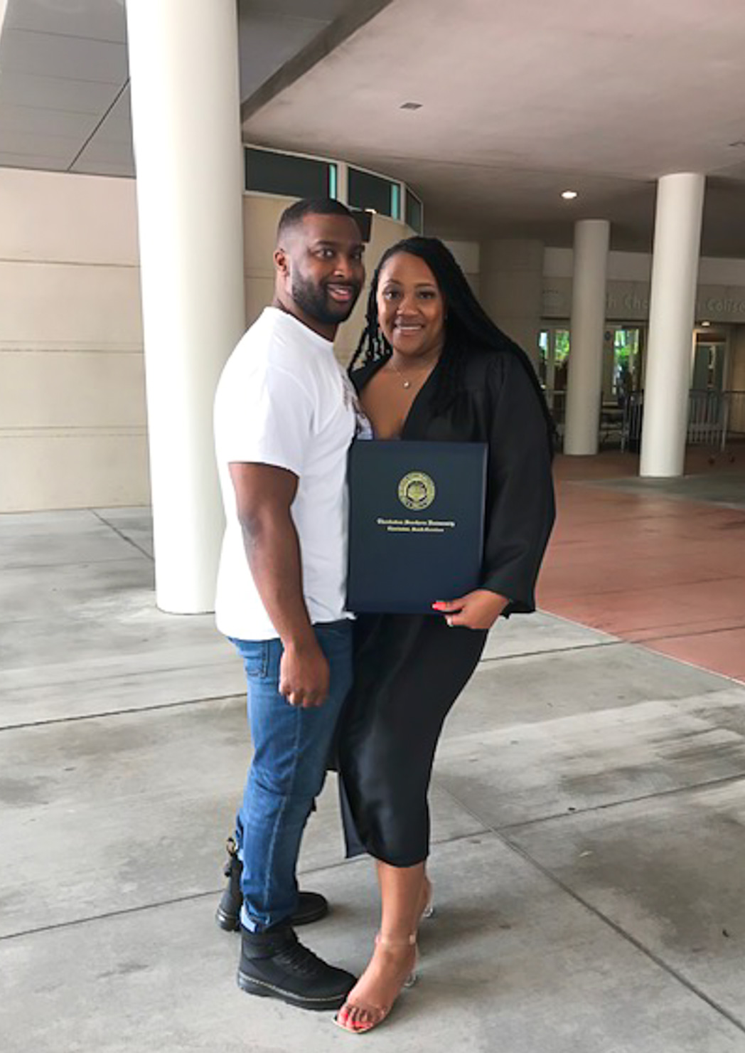 A man in a white T-shirt and jeans stands beside a smiling woman in a black graduation gown holding a navy diploma cover with a gold seal, under a large concrete overhang. He has his arm around her, and she holds the folder in front of her with both hands. The cover has a gold seal and gold lettering that appears to read "Charleston Southern University," They stand on gray pavement, with white columns and a building entrance behind them.