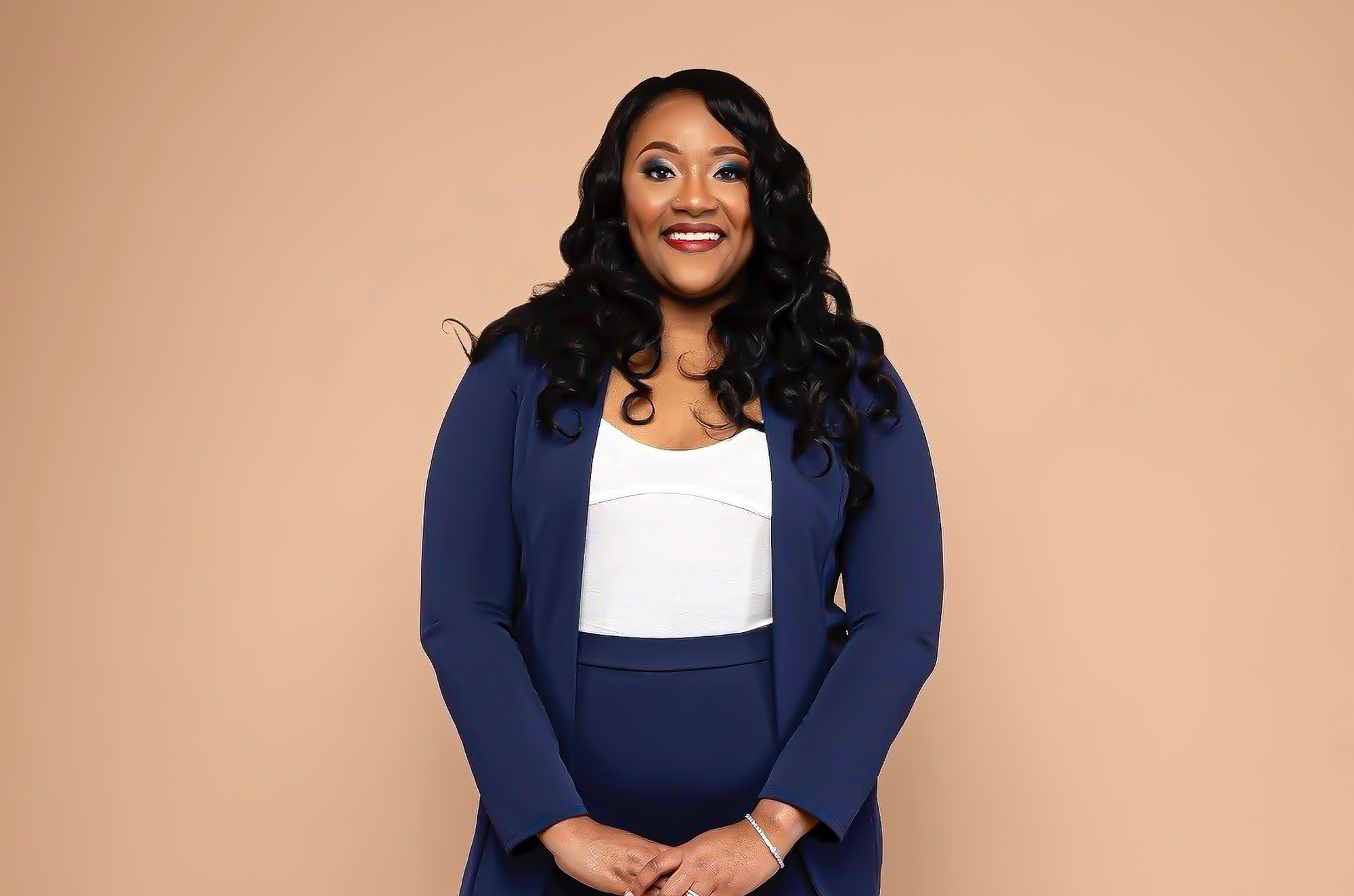 Studio portrait of a smiling woman with long, black, loosely curled hair, wearing a navy blazer over a white top and navy skirt, hands clasped in front of a plain warm peach backdrop. She wears blue eyeshadow, red lipstick, and a sparkling bracelet on one wrist, and she faces the camera directly with a warm, confident smile.