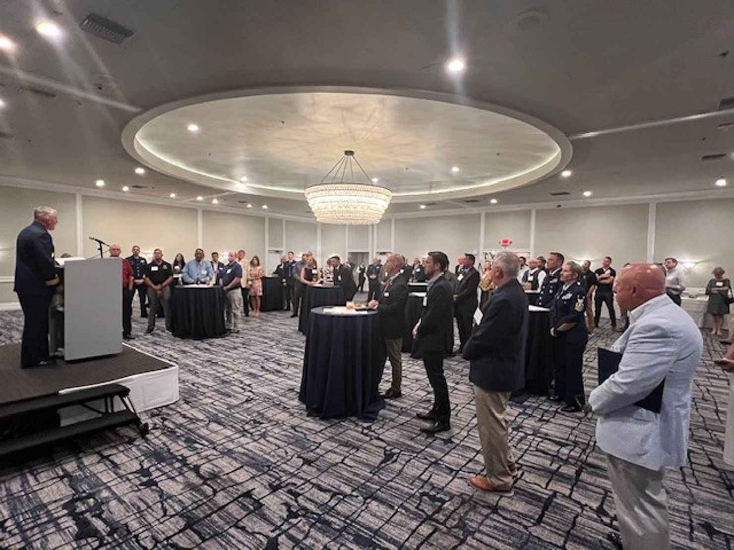 A speaker in dark uniform addresses a ballroom of standing guests gathered around cocktail tables, under a large round chandelier and domed ceiling.