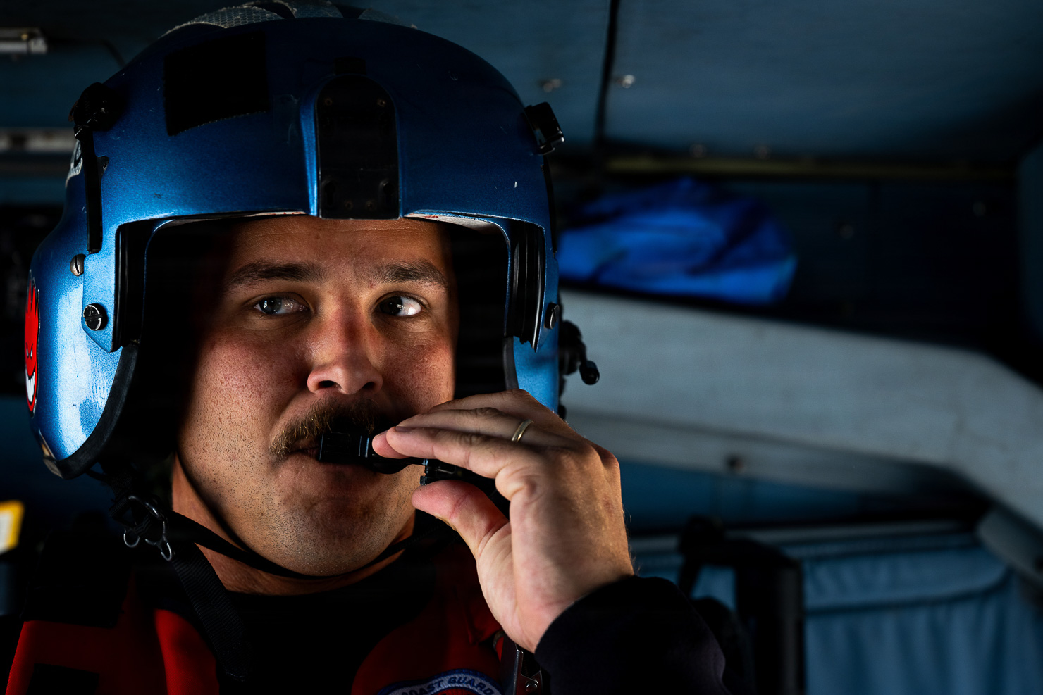 Close-up portrait of a man in a blue flight helmet speaking into a boom microphone inside an aircraft cabin.