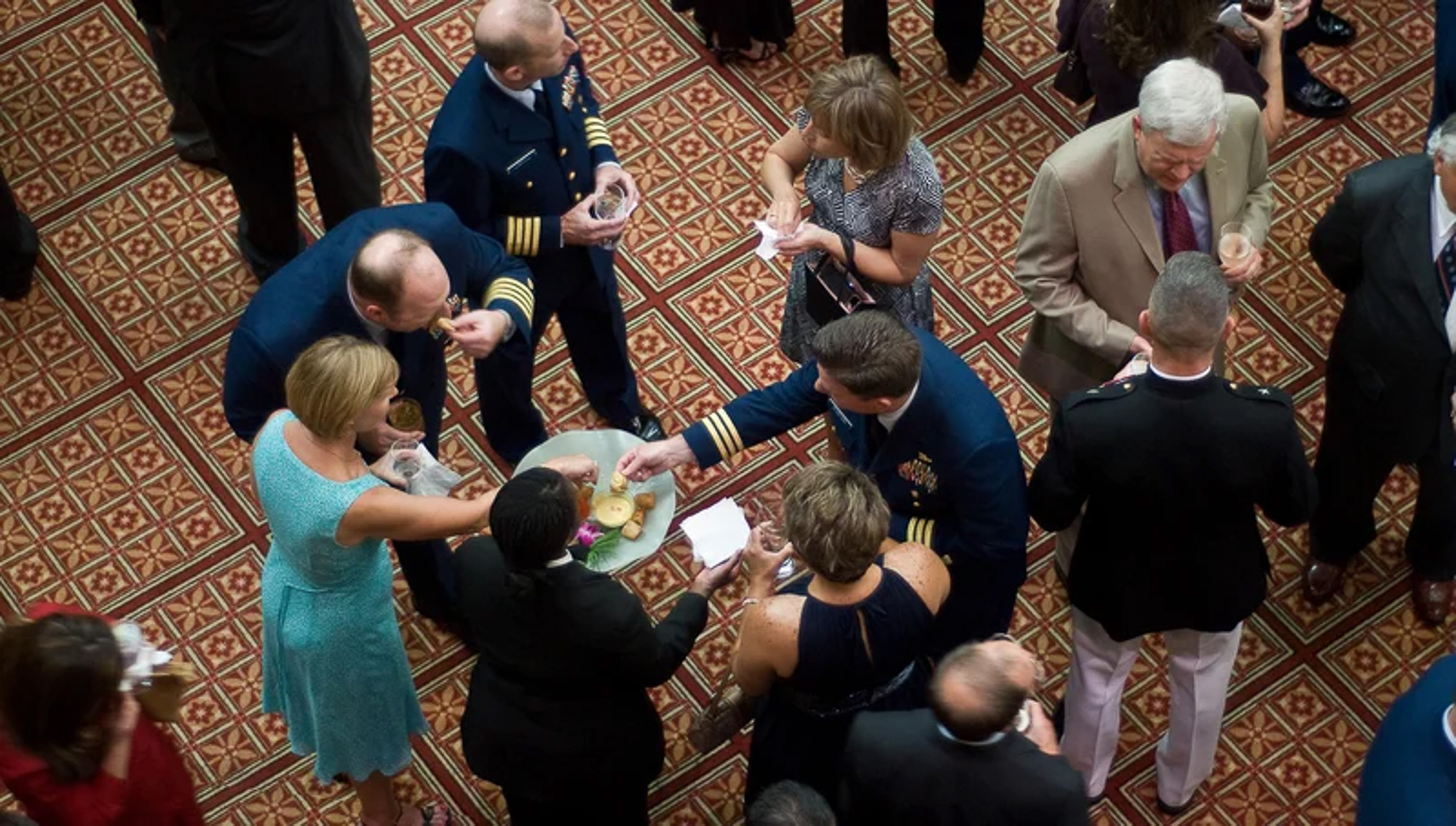 Overhead view of guests in formal and military dress uniforms mingling around a shared appetizer plate on a patterned carpet.