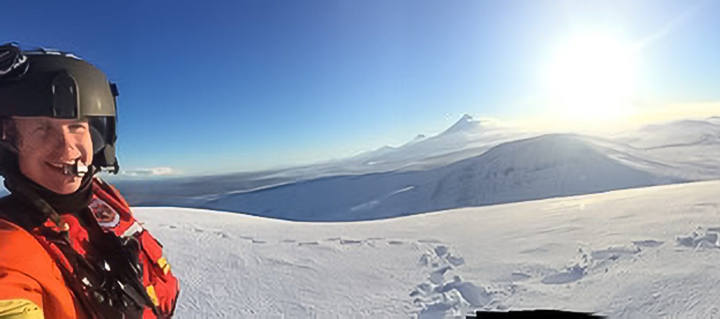 Man takes a selfie in the Arctic, showing the snow-covered mountains and sun beyond the peaks in the distance. The sky is clear and blue.