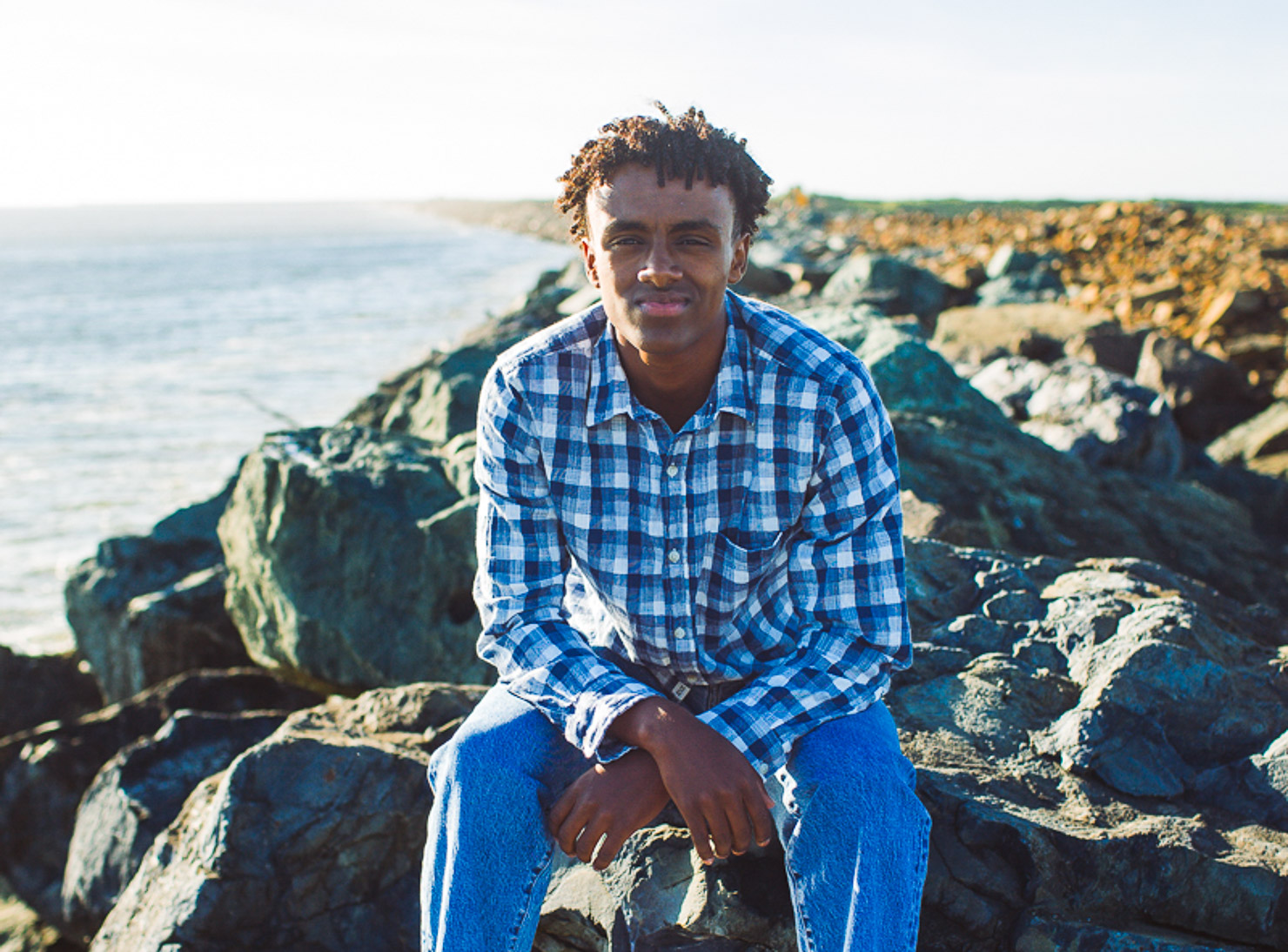 Boy sits on a large rock on a rocky beach overlooking the Pacific Ocean. He's dressed in a long sleeve, blue-checkered button down shirt. His posture is relaxed as he sits forward with his elbows resting on his knees and wrists crossed one over the other. He's smiling and slightly squinting as the sunlight illuminates one side of his face.