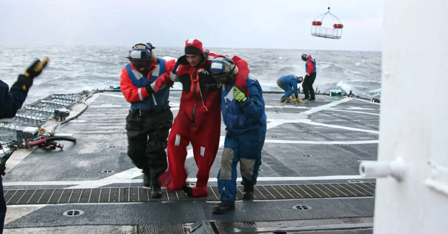 Two men hold a third man up between them, helping him as he walks on a ship in the middle of the ocean. He has just been rescued from the freezing waters of the Arctic Ocean.