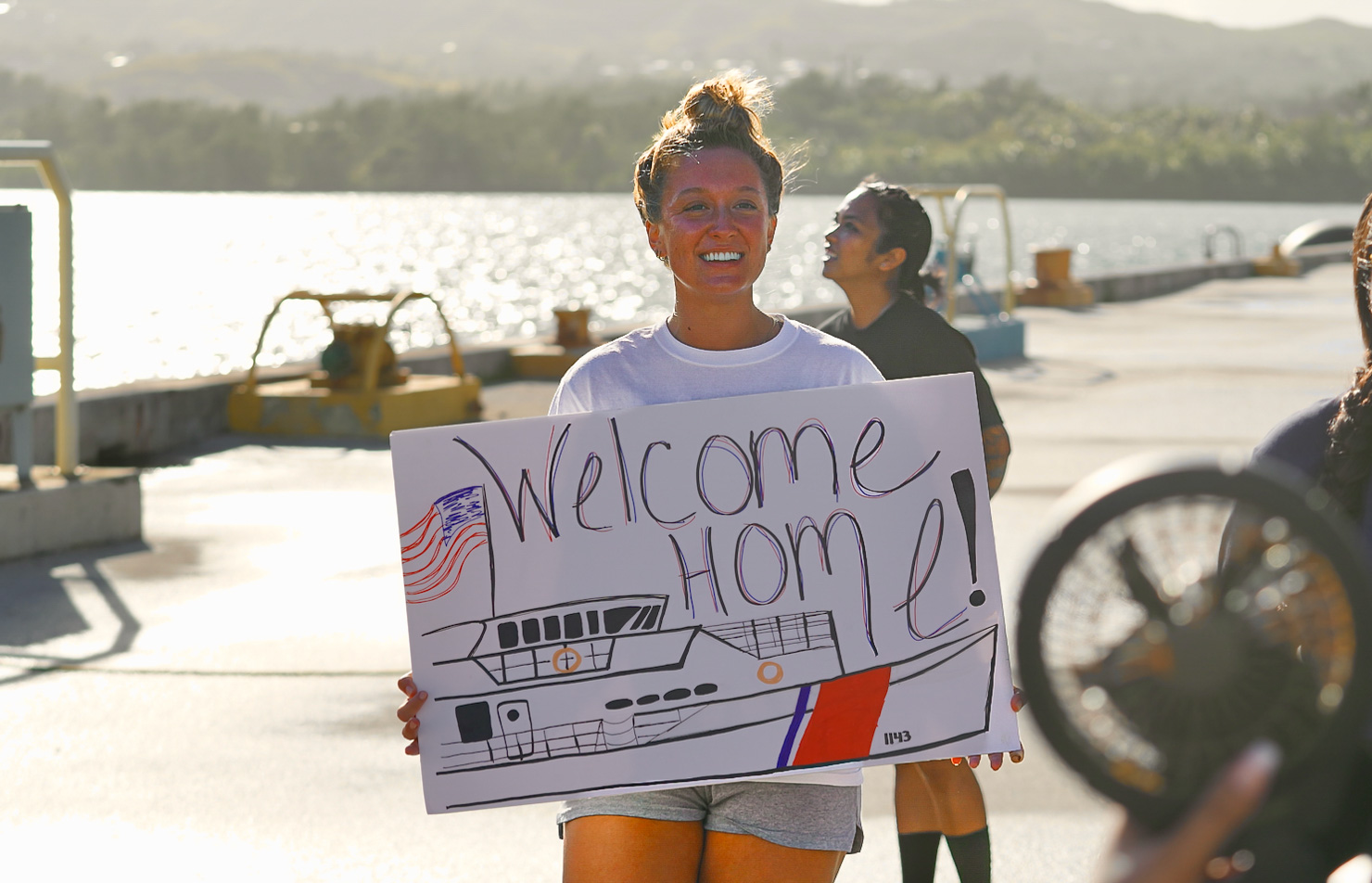 Blond woman stands on a dock, holding a "welcome home" sign while smiling.