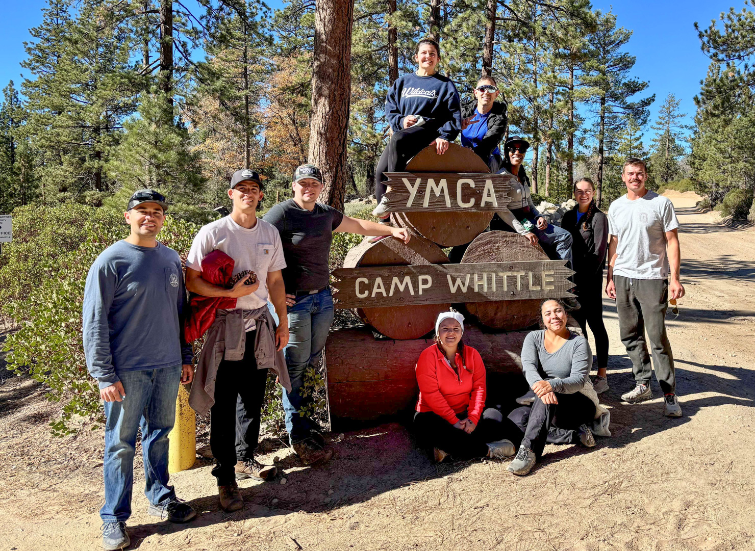 Group poses for a photo in the middle of the woods, around a large rustic sign that reads "YMCA. Camp Whittle".