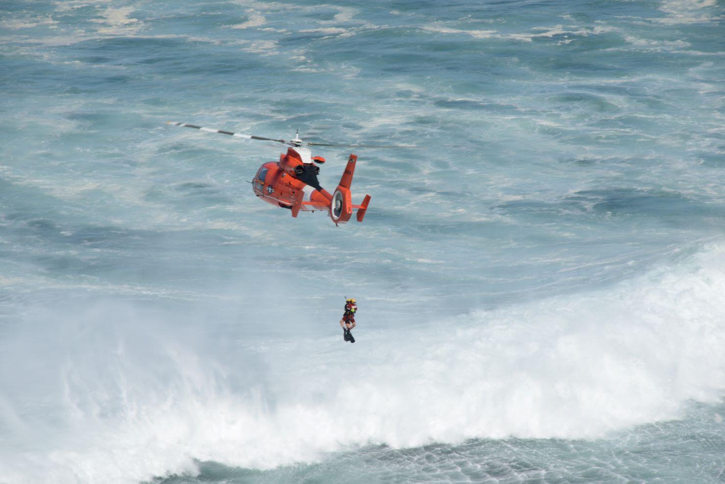 A Coast Guard helicopter hovering over roiling surf hosting a rescue swimmer.