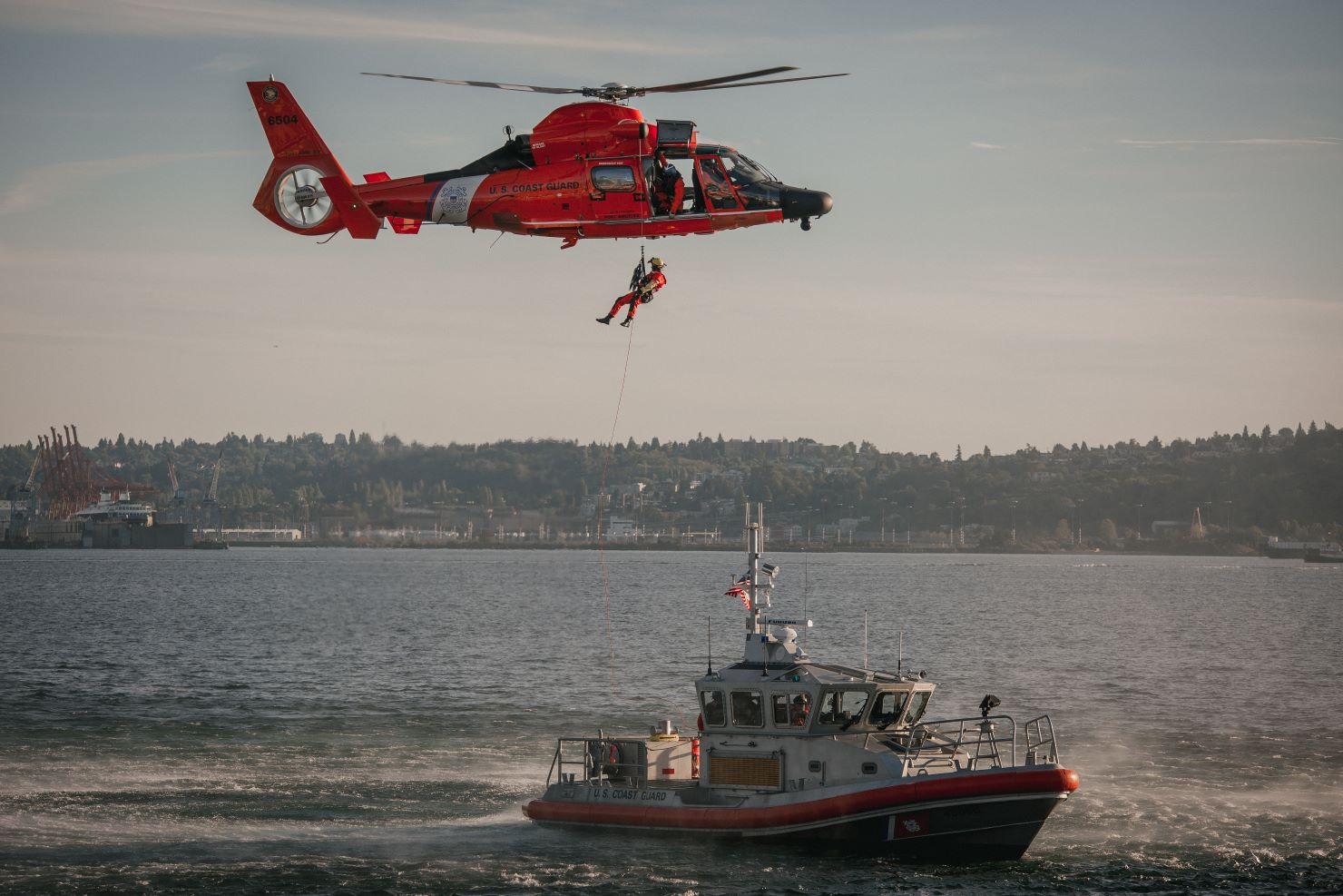 A Coast Guard helicopter hovers over a Coast Guard boat while hoisting a rescue swimmer